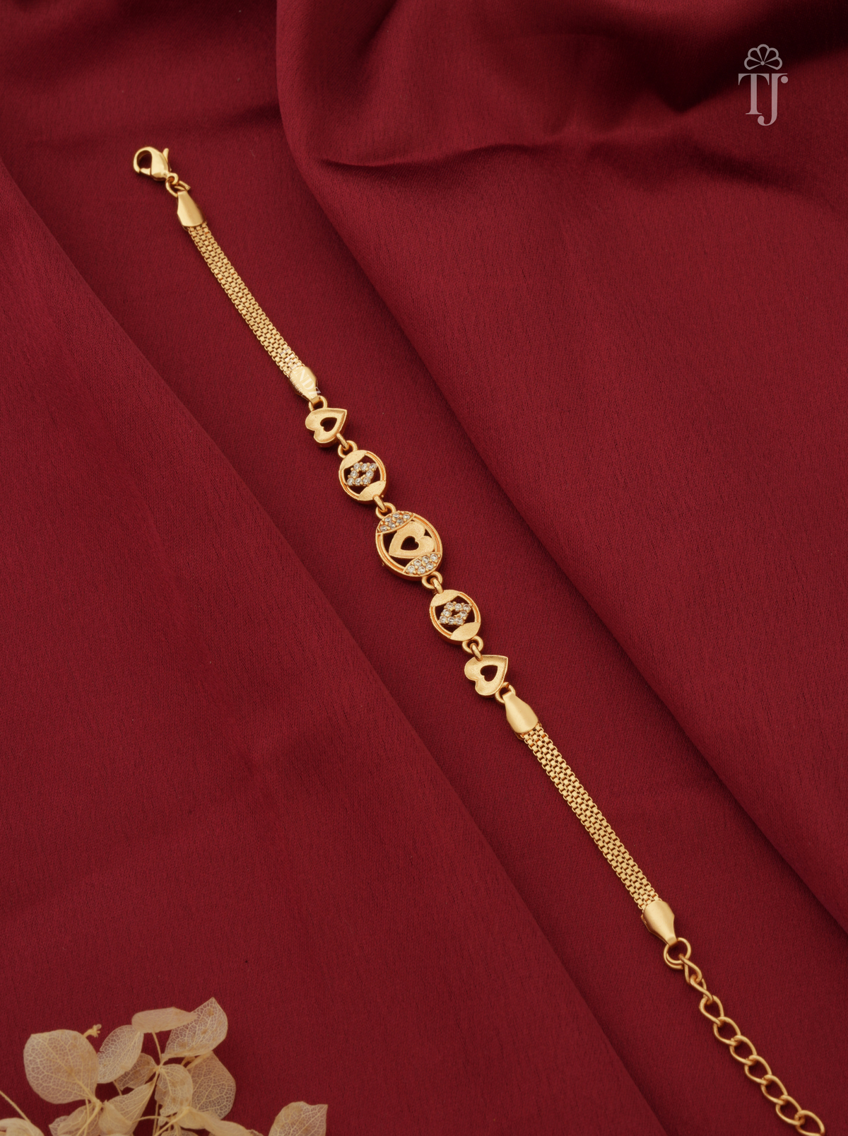 A gold-plated chain bracelet with circular and floral motifs, laid diagonally on draped dark red fabric.