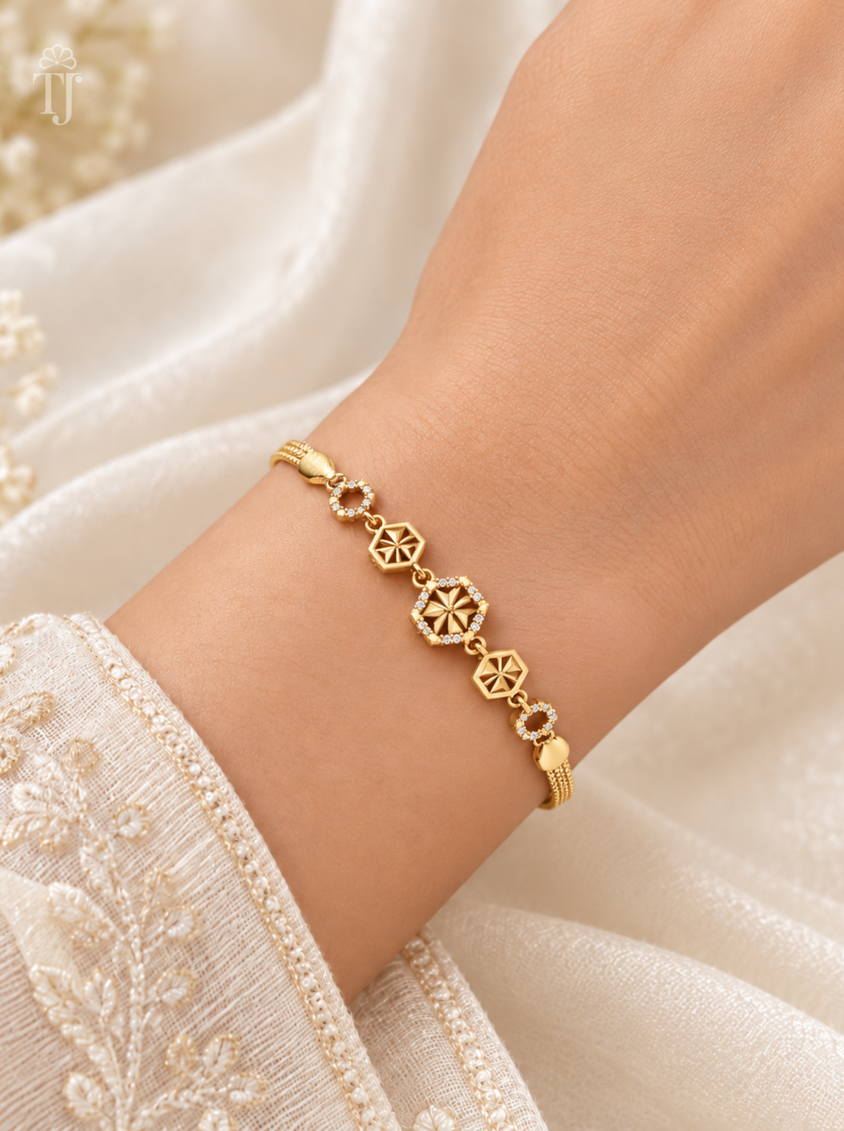 A delicate gold bracelet with geometric and floral motifs worn on a person's wrist, set against a soft cream-colored embroidered fabric background.