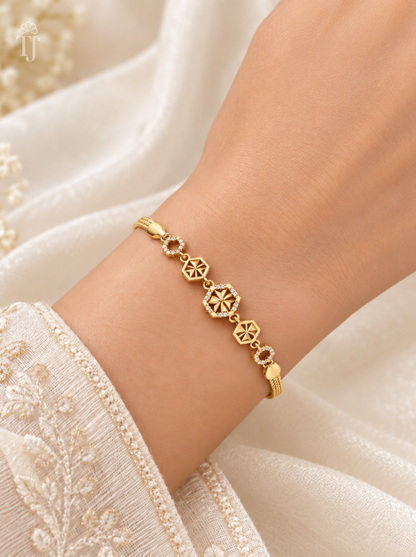 A delicate gold bracelet with geometric and floral motifs worn on a person's wrist, set against a soft cream-colored embroidered fabric background.