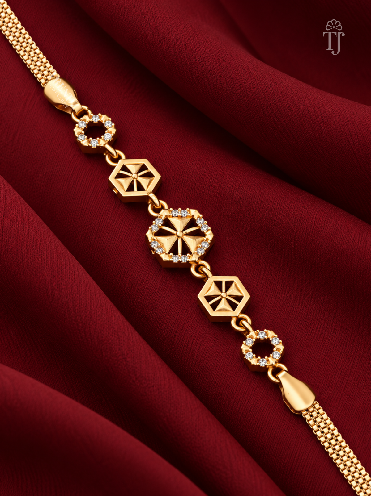 A gold-toned chain bracelet with geometric star and circular link motifs, some accented with small clear stones, displayed on draped deep red fabric.