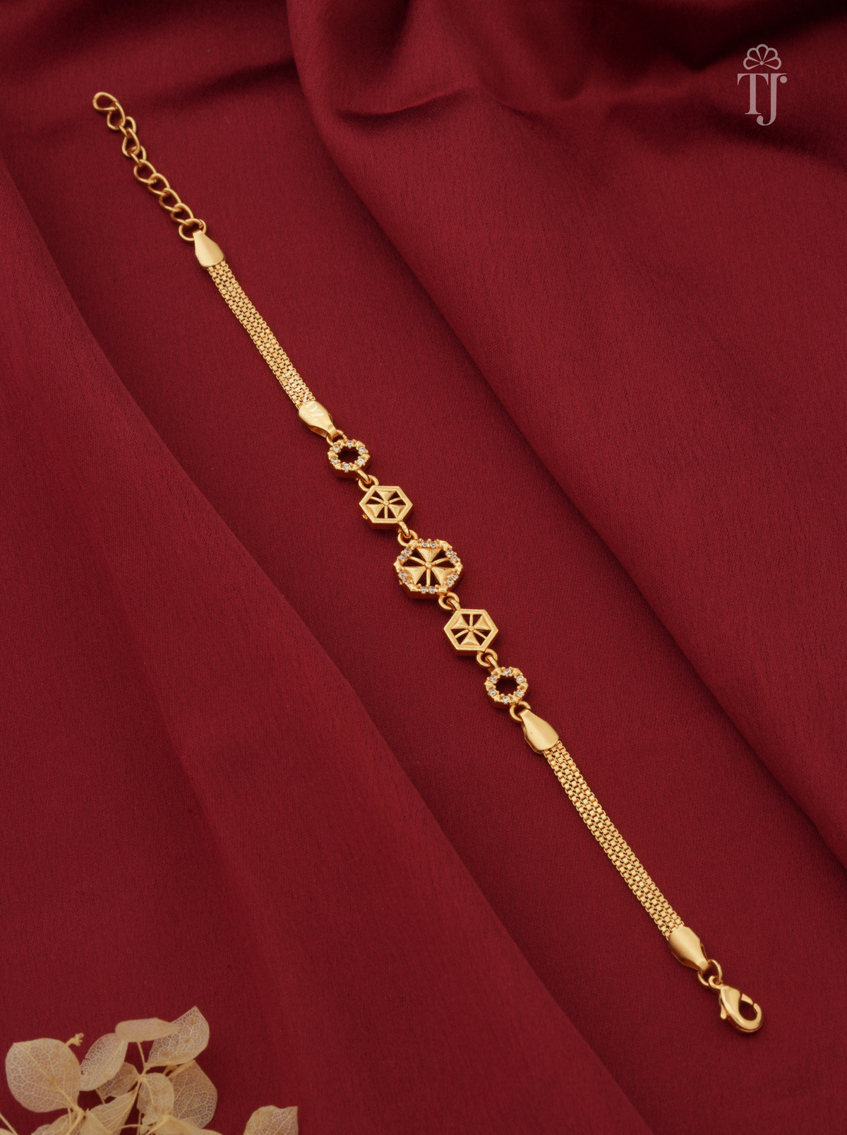 A delicate gold chain bracelet with ornate geometric and floral motifs, displayed diagonally on a draped dark red fabric background.