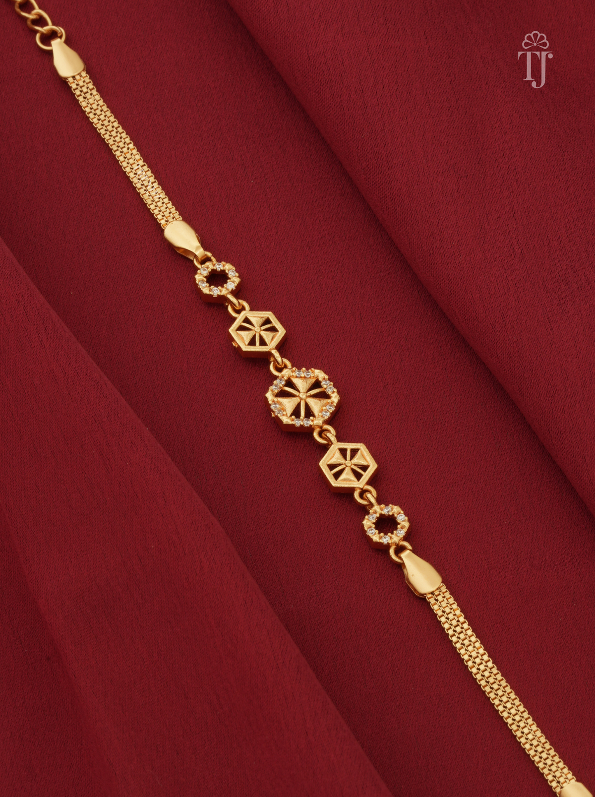 A gold-toned bracelet with geometric star and circular link motifs, displayed diagonally on draped dark red fabric.