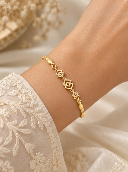 A delicate gold bracelet with geometric diamond-shaped links and a small central motif, shown worn on a person's wrist. The person is wearing a cream-colored embroidered sleeve, with a soft, draped cream fabric background.