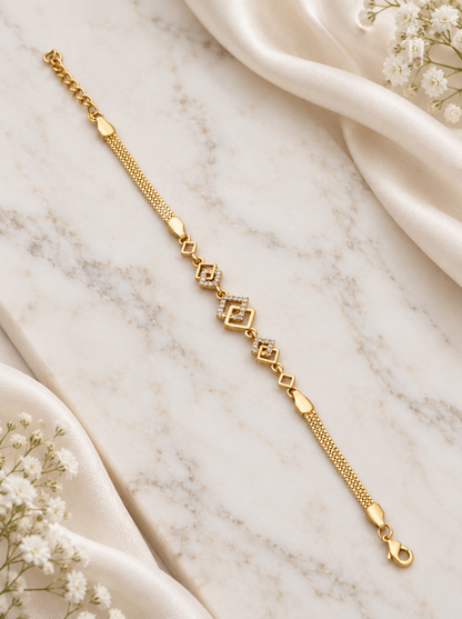 A delicate gold chain bracelet with geometric diamond-shaped links and heart-shaped accents, displayed on a white marble surface with draped white satin and small white flowers.