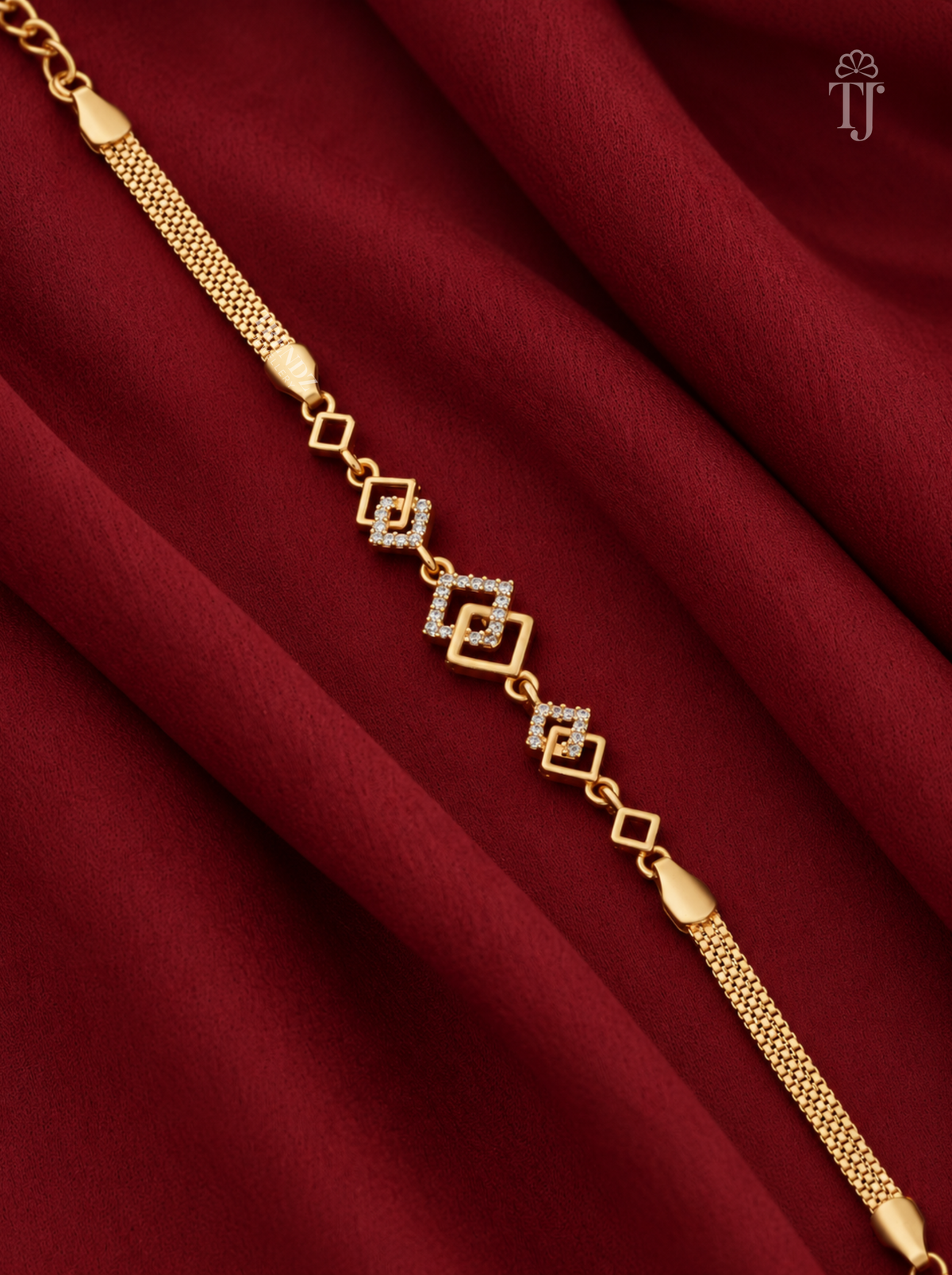 A gold chain bracelet with a central geometric diamond motif set with small clear stones, displayed diagonally on draped dark red fabric. A small 'TJ' logo watermark is visible in the top right corner.