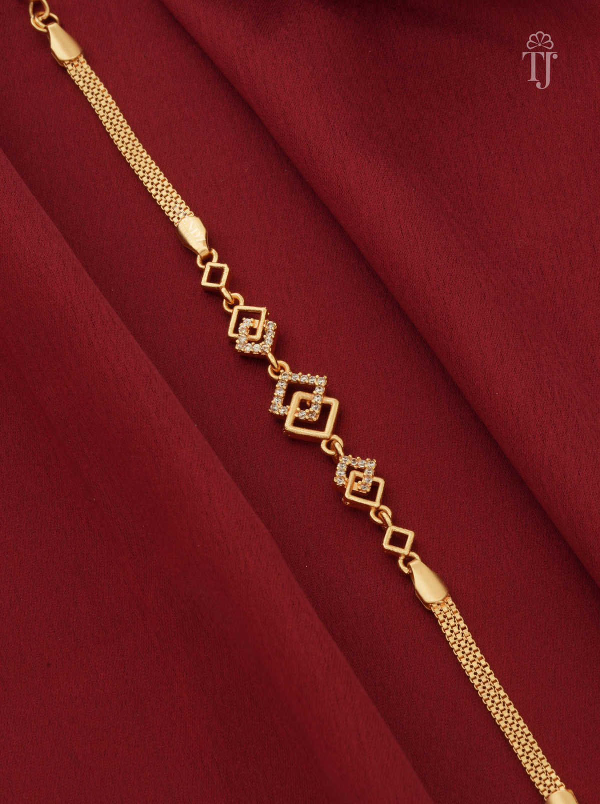 A gold chain bracelet with a central diamond-shaped motif paved with small clear stones, displayed diagonally on draped dark red fabric.