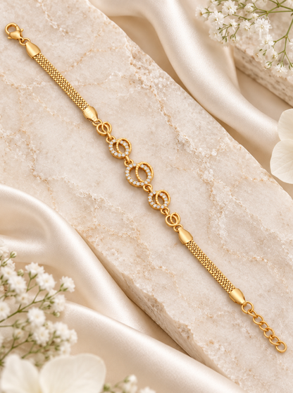 A gold chain bracelet with three oval links set with small clear stones in the center, displayed on a textured cream stone slab with draped satin fabric and white flowers.