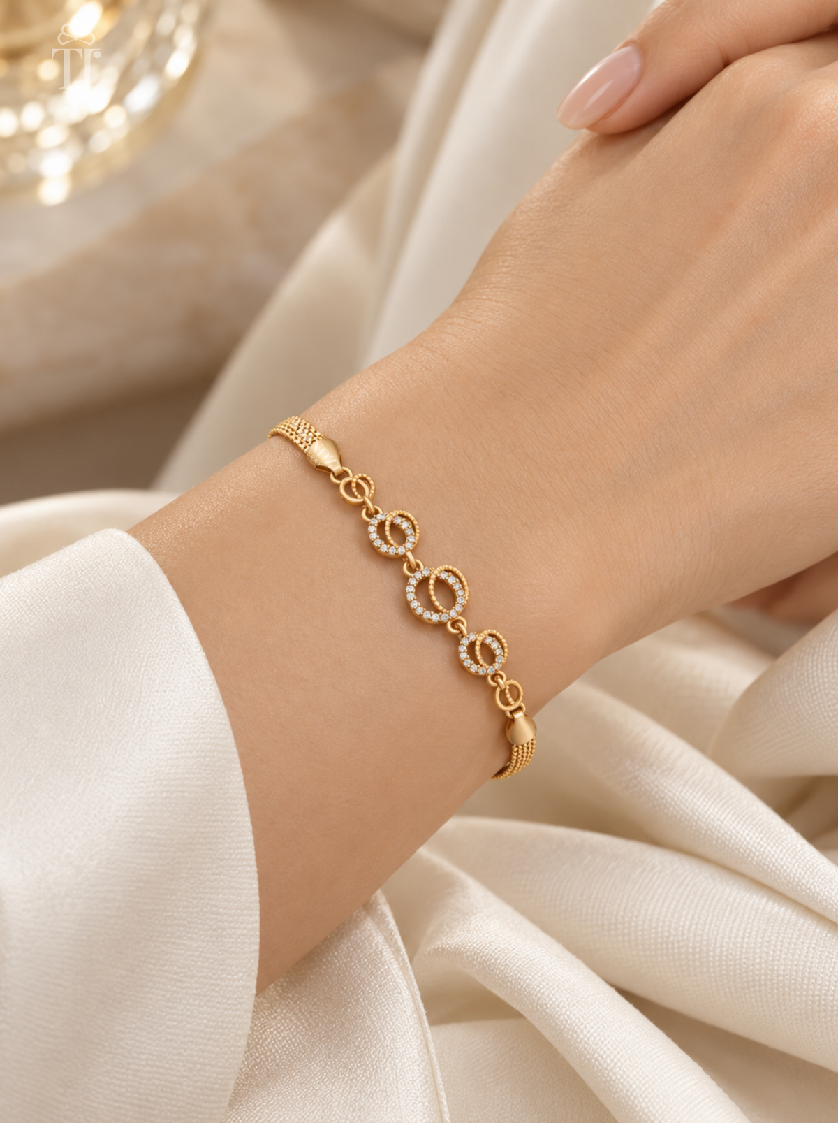 A delicate gold bracelet with interlocking circular links, some adorned with small clear stones, worn on a woman's wrist. The background consists of soft, draped white fabric.