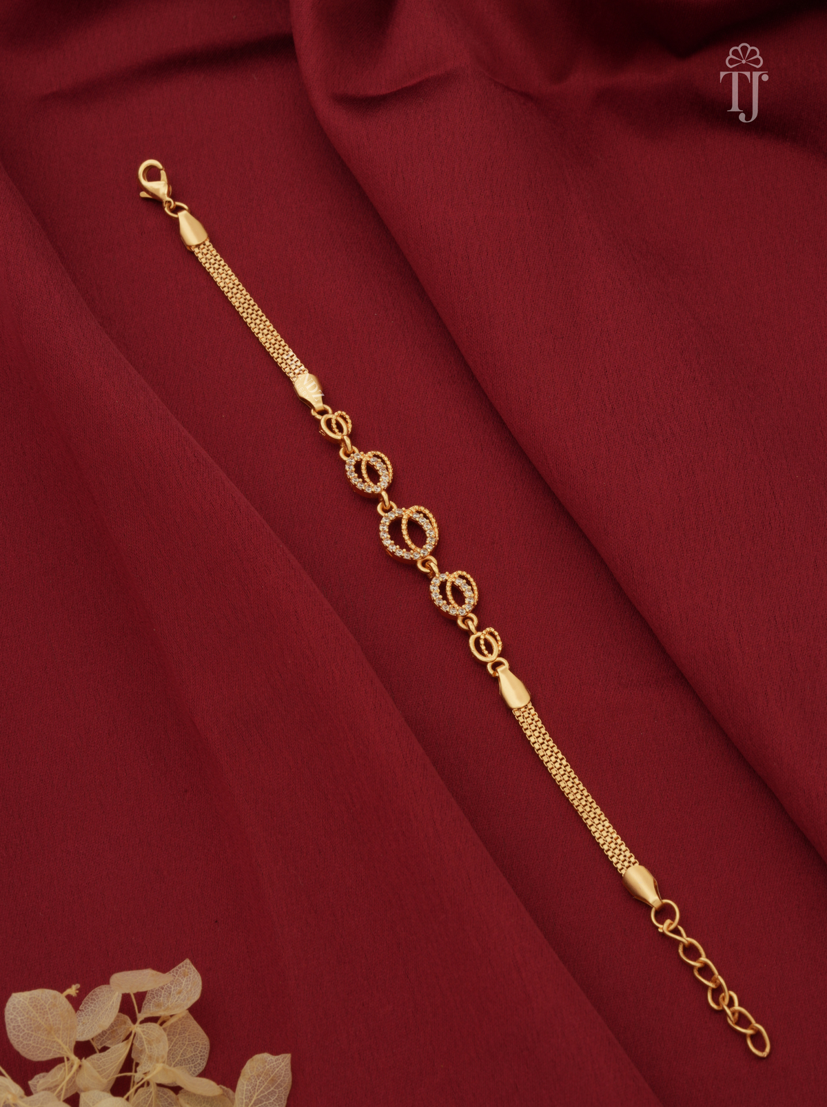 A gold-toned chain bracelet with alternating oval and circular links, displayed diagonally on draped dark red fabric with a small dried floral accent in the corner.