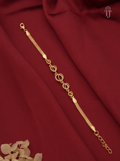 A gold-toned chain bracelet with alternating oval and circular links, displayed diagonally on draped dark red fabric with a small dried floral accent in the corner.