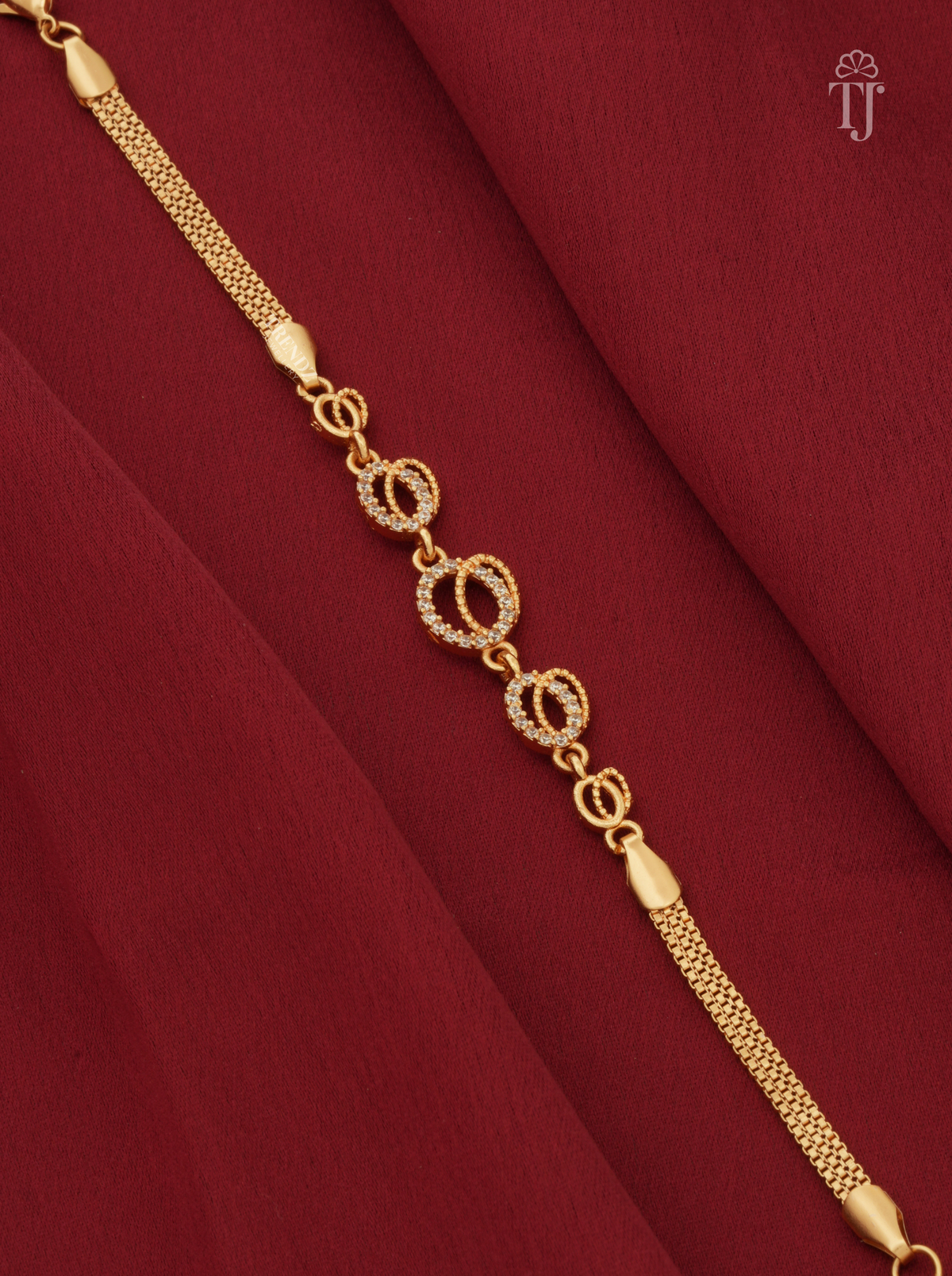 A gold chain bracelet with three oval links encrusted with small clear stones, displayed diagonally on draped deep red fabric.