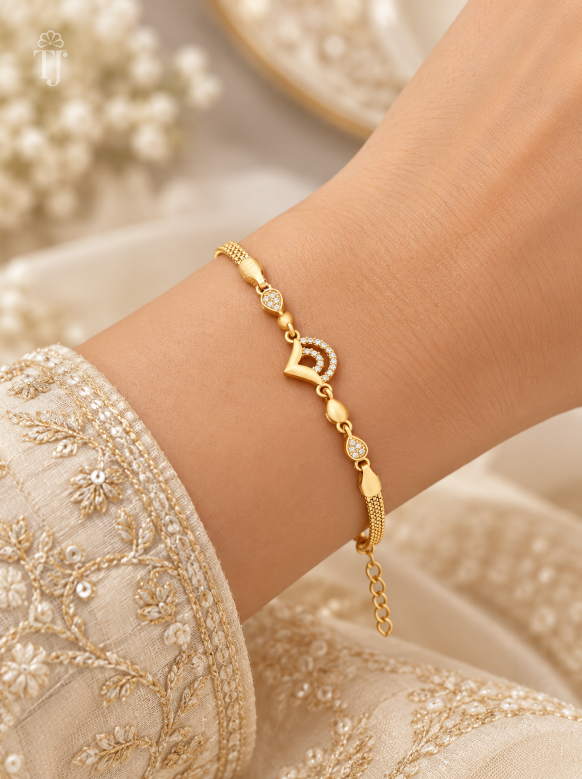 A delicate gold chain bracelet with a small heart-shaped charm and tiny clear stones, worn on a person's wrist. The person is wearing a cream-colored embroidered traditional garment, with a soft, blurred background of similar cream tones.