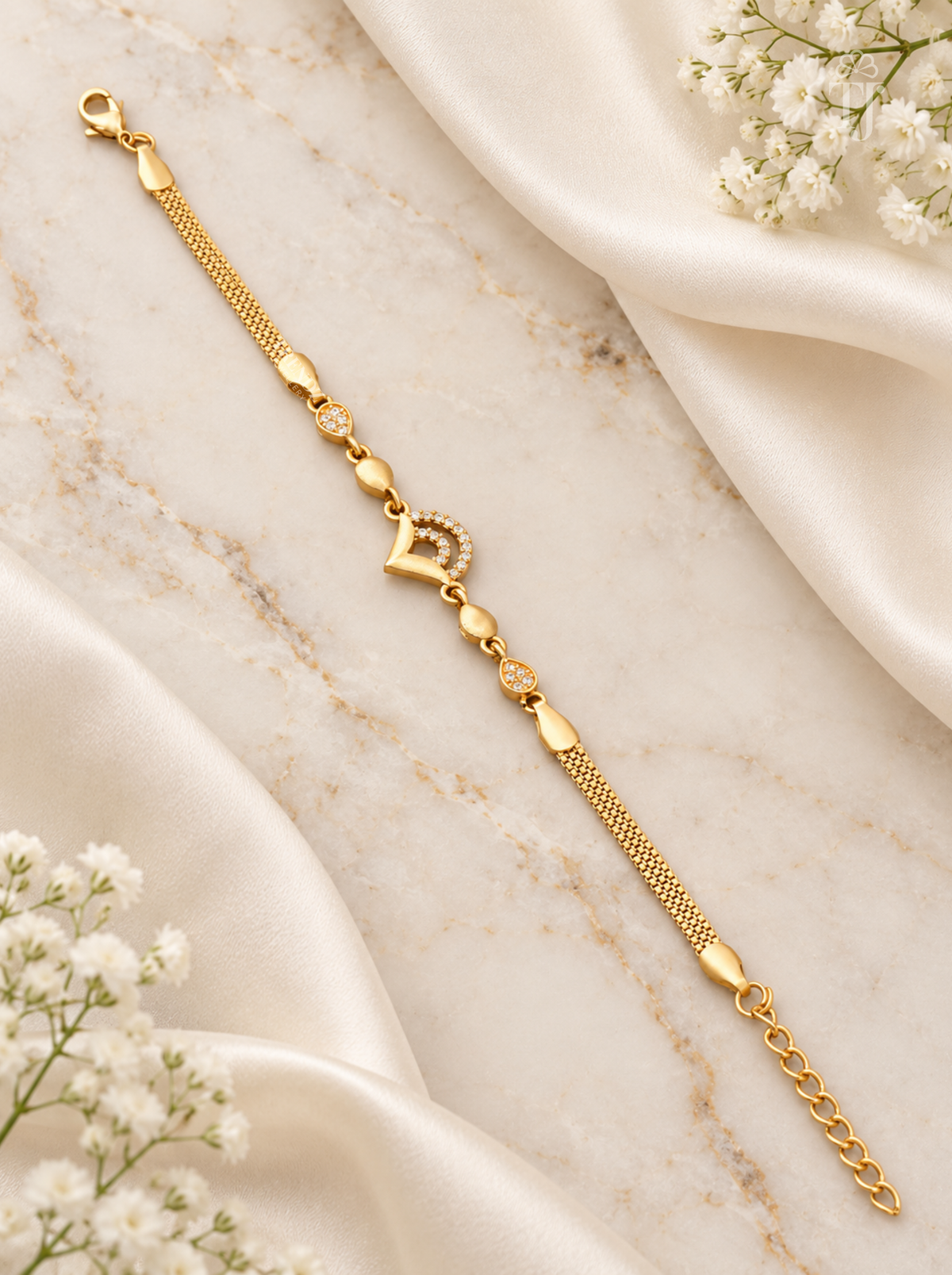 A delicate gold chain bracelet with a central heart-shaped charm and small clear stones, styled on a cream marble surface with draped satin and white baby's breath flowers.