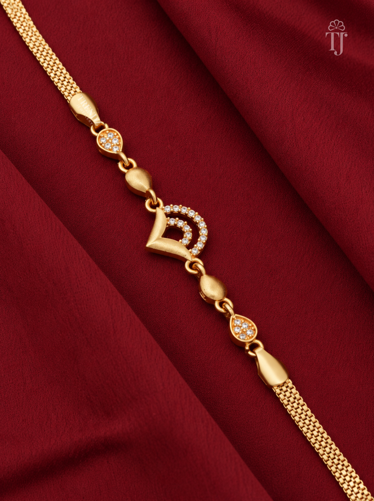 A gold chain bracelet with a central crescent moon and heart motif embellished with small clear stones, displayed diagonally on a draped dark red fabric background.