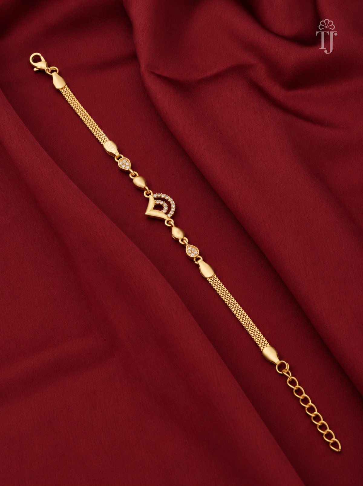 A gold-plated chain bracelet with a central heart-shaped charm and small decorative beads, displayed diagonally on a draped dark red fabric background.