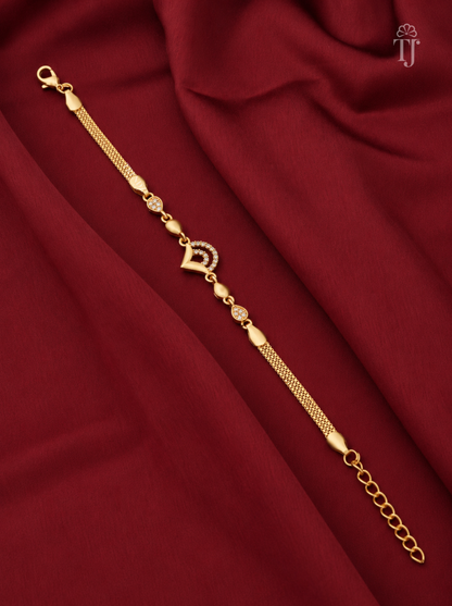 A gold-plated chain bracelet with a central heart-shaped charm and small decorative beads, displayed diagonally on a draped dark red fabric background.