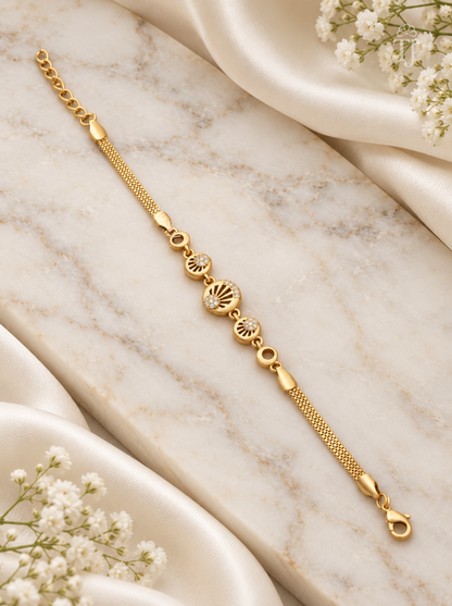 A gold chain bracelet with three decorative oval charms, laid diagonally on a white marble slab with white satin fabric and small white flowers as props.