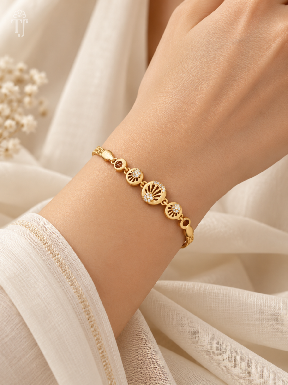 A delicate gold bracelet with three sunburst wheel charms and small clear stones, shown worn on a person's wrist against a soft cream fabric background.