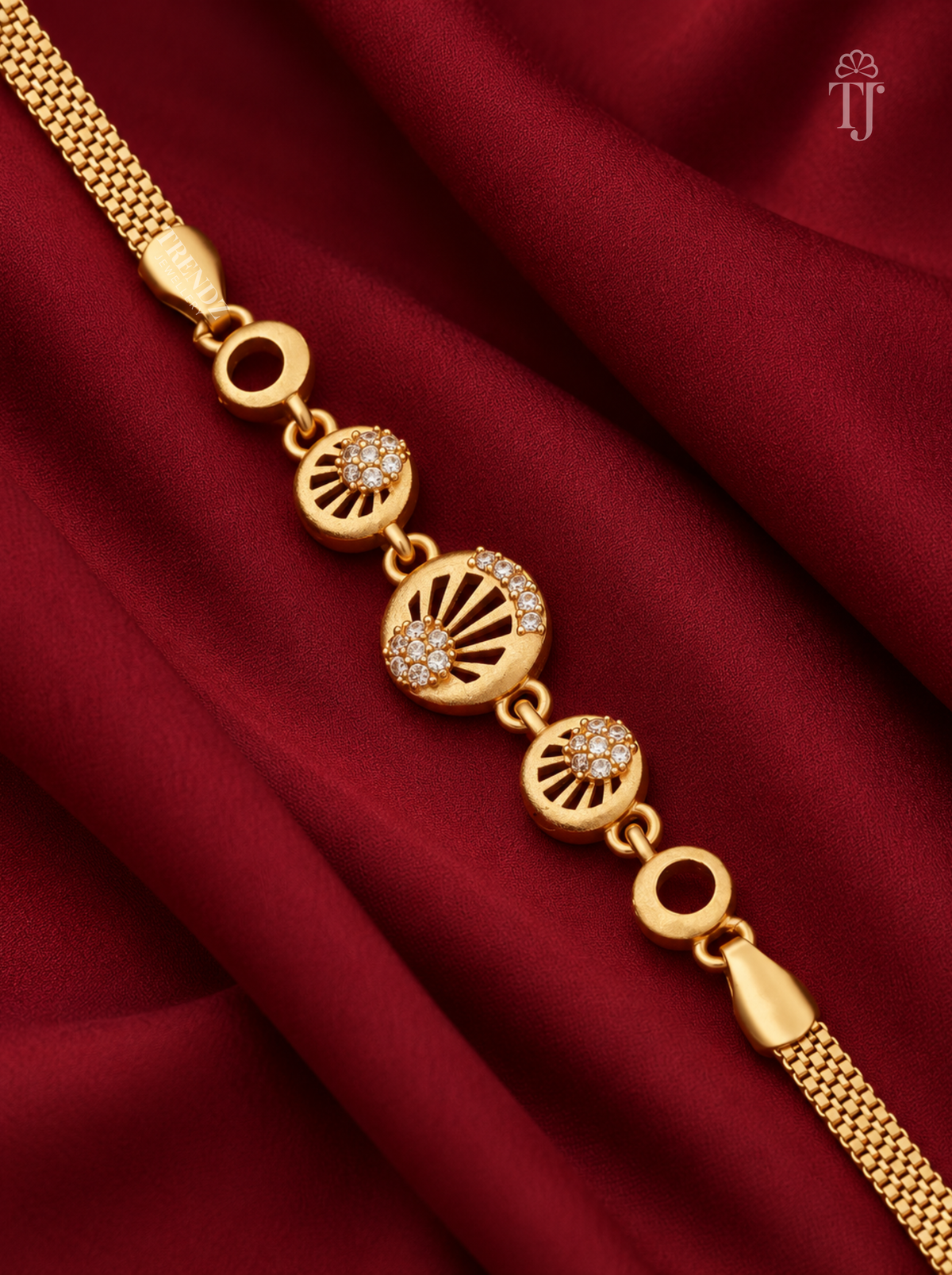 A gold chain bracelet with three circular fan-shaped charms, two of which are adorned with small clear stones, displayed on a draped dark red satin fabric. A small 'TJ' logo is visible in the top right corner.