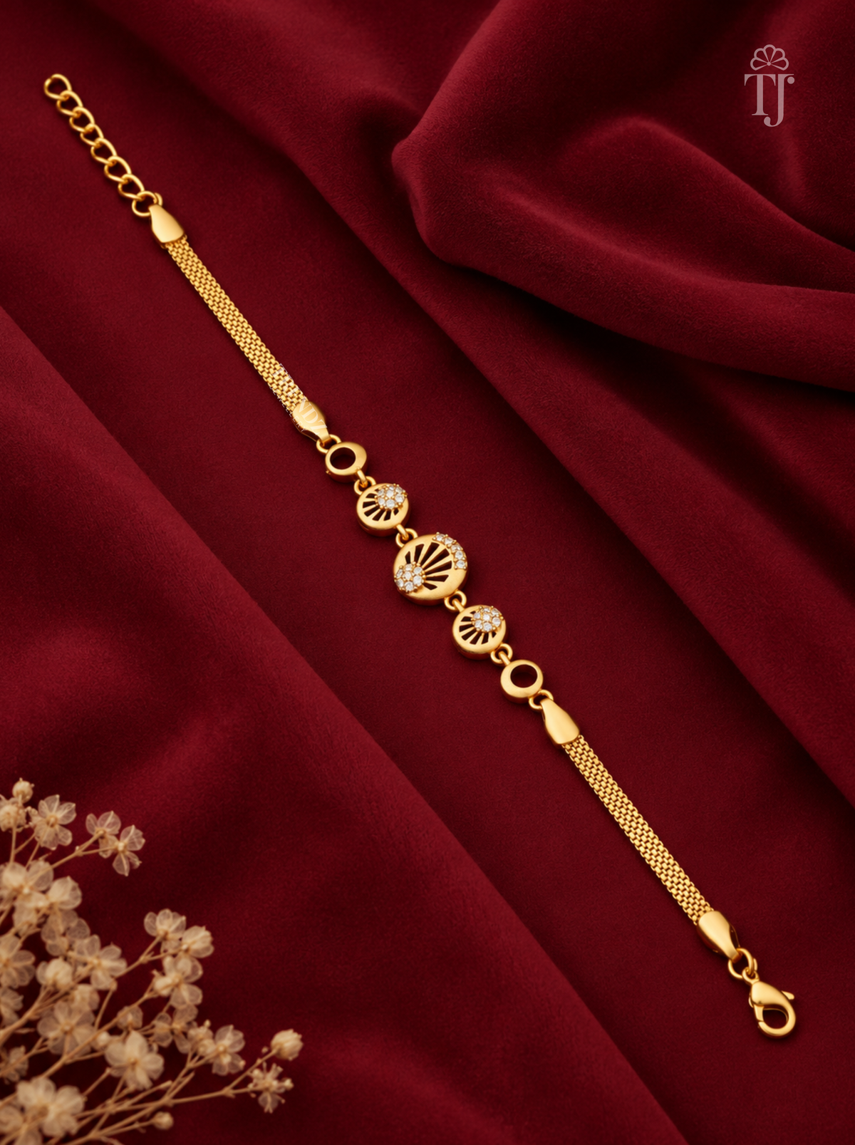 A gold-plated chain bracelet with three decorative filigree and oval links, displayed diagonally on a draped deep red velvet fabric background with dried flowers in the corner.