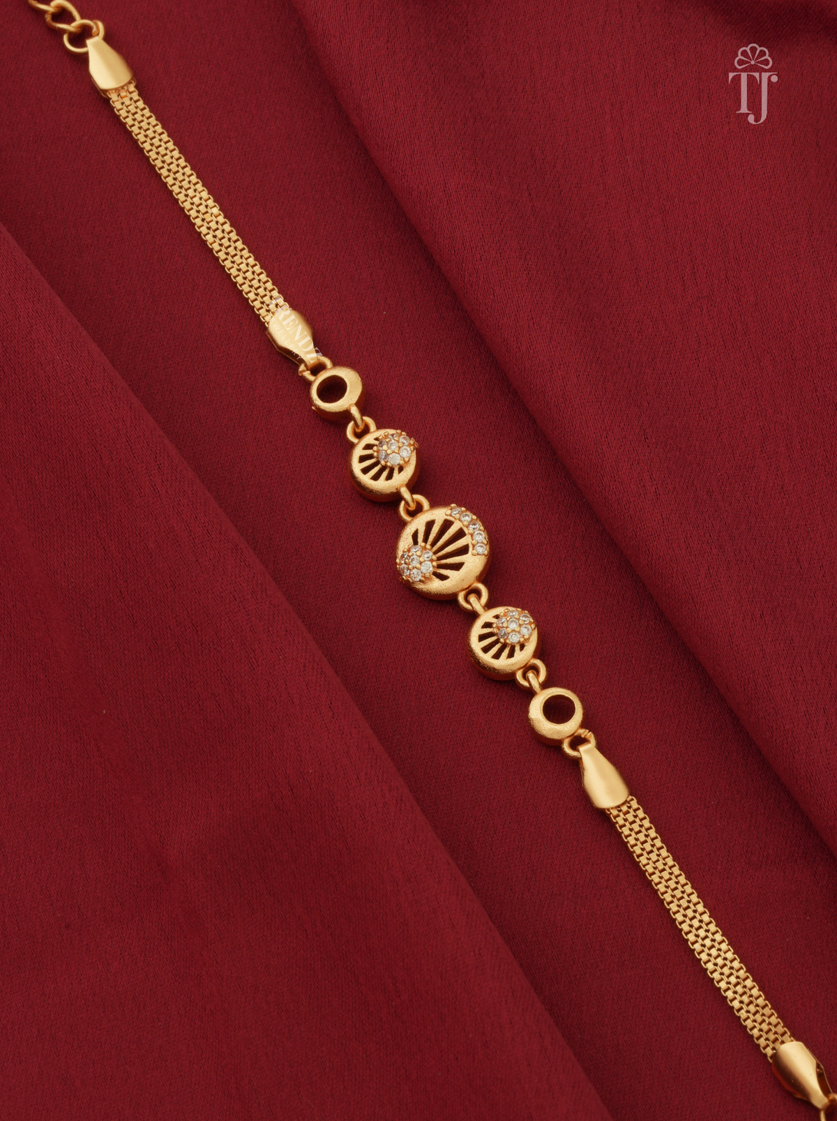 A gold chain bracelet with three circular decorative charms, two of which feature small clear stones, displayed diagonally on a draped dark red fabric background.