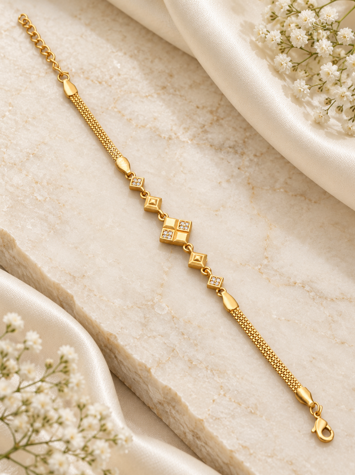 A gold chain bracelet with small diamond-shaped links, some set with clear stones, displayed on a light beige stone slab with white fabric and baby's breath flowers.