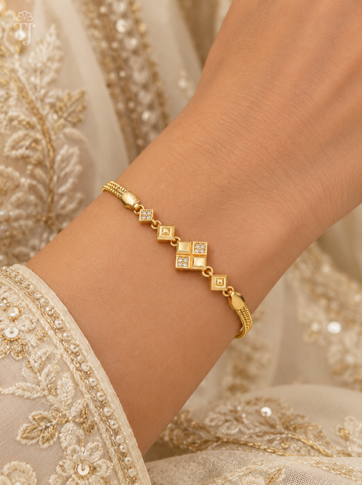 A delicate gold bracelet with diamond-shaped motifs and small clear stones, shown worn on a person's wrist against an ornate cream-colored embroidered garment.