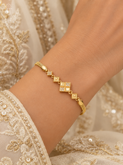 A delicate gold bracelet with diamond-shaped motifs and small clear stones, shown worn on a person's wrist against an ornate cream-colored embroidered garment.