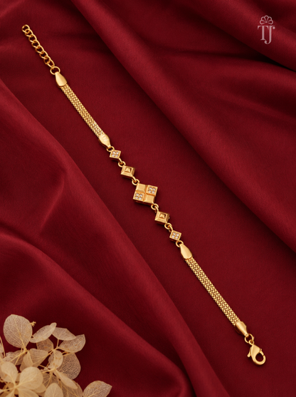 A gold chain bracelet with a central diamond-shaped charm and smaller diamond accents, displayed on a draped dark red satin fabric background with dried flowers in the corner.