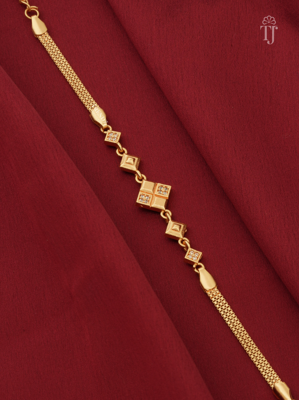 A gold chain anklet with a central diamond-shaped charm set with small clear stones, displayed diagonally on a draped dark red fabric background.