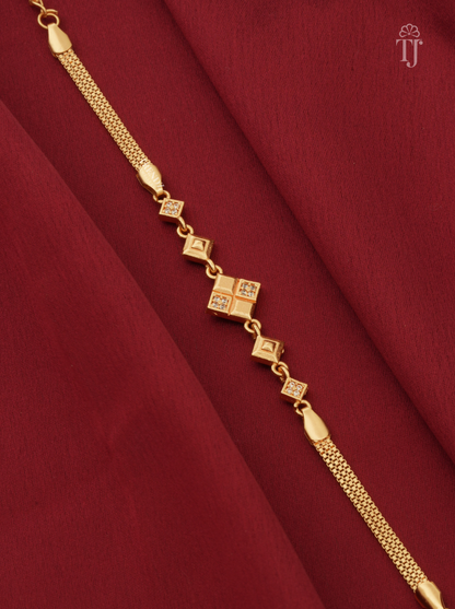A gold chain anklet with a central diamond-shaped charm set with small clear stones, displayed diagonally on a draped dark red fabric background.