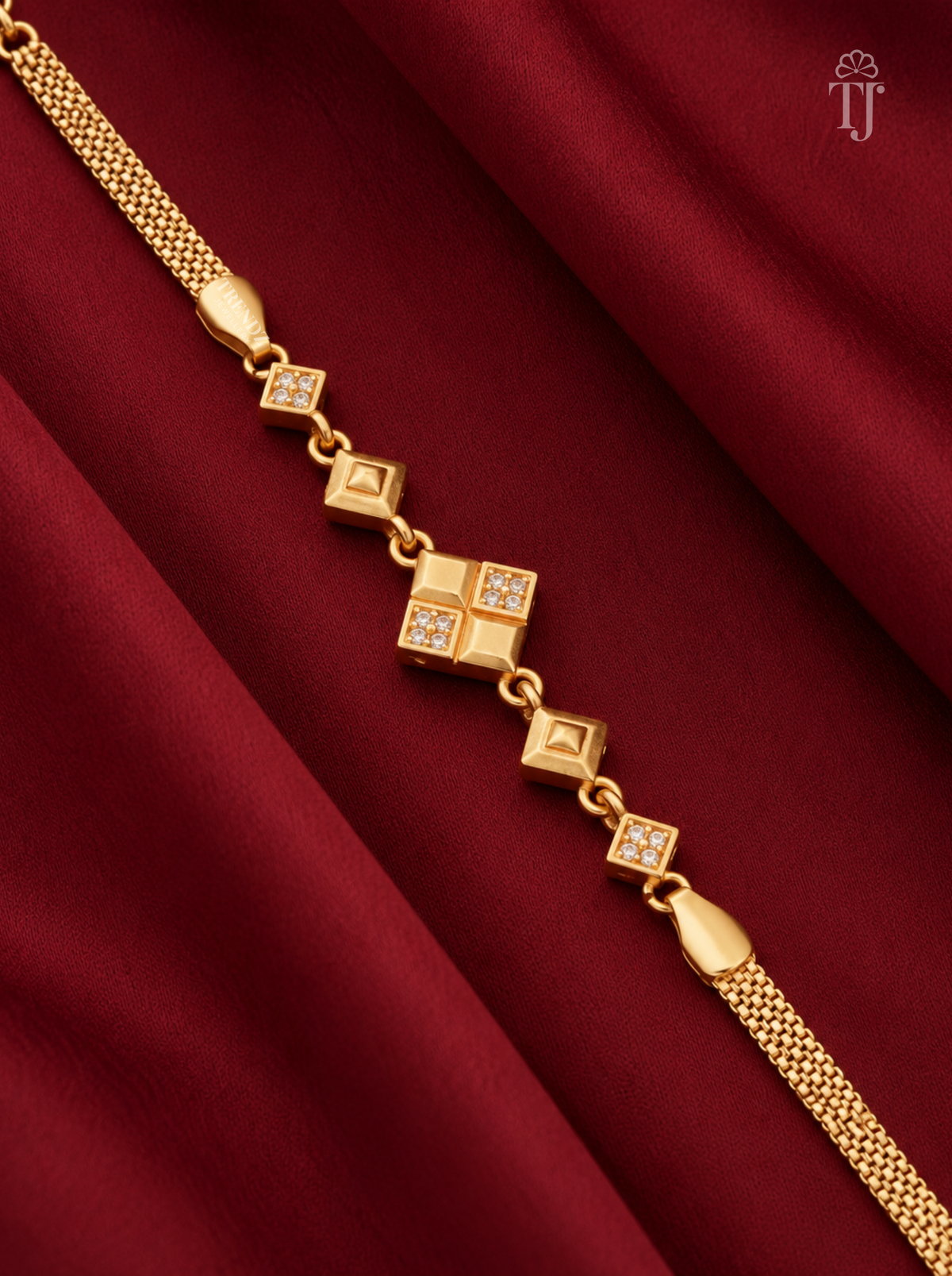 A gold chain bracelet with alternating square links, some adorned with small clear stones, displayed diagonally on a draped dark red fabric background.