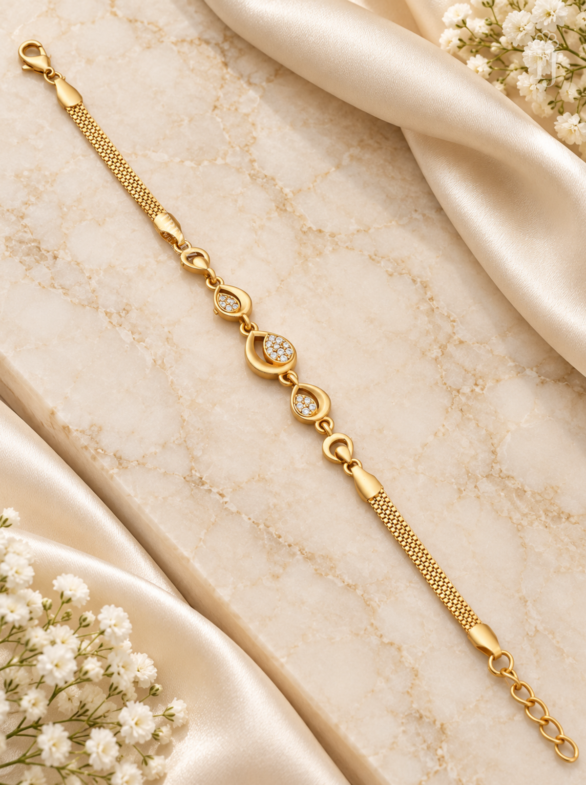 A gold chain bracelet with three oval links set with small clear stones, displayed on a beige marble surface with cream satin fabric and white baby's breath flowers.