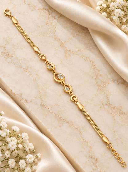 A gold chain bracelet with three oval links set with small clear stones, displayed on a beige marble surface with cream satin fabric and white baby's breath flowers.