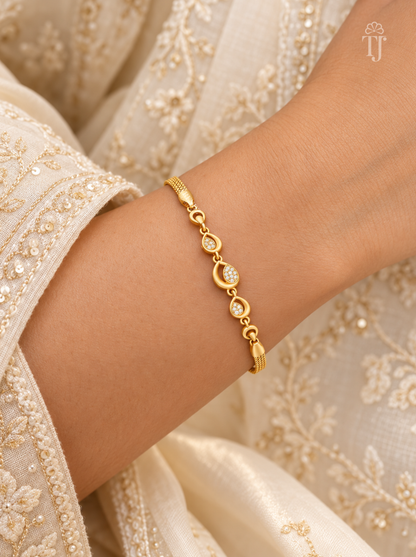 A delicate gold chain bracelet with small circular links and clear stones, worn on a person's wrist. The person is wearing an off-white embroidered traditional garment.