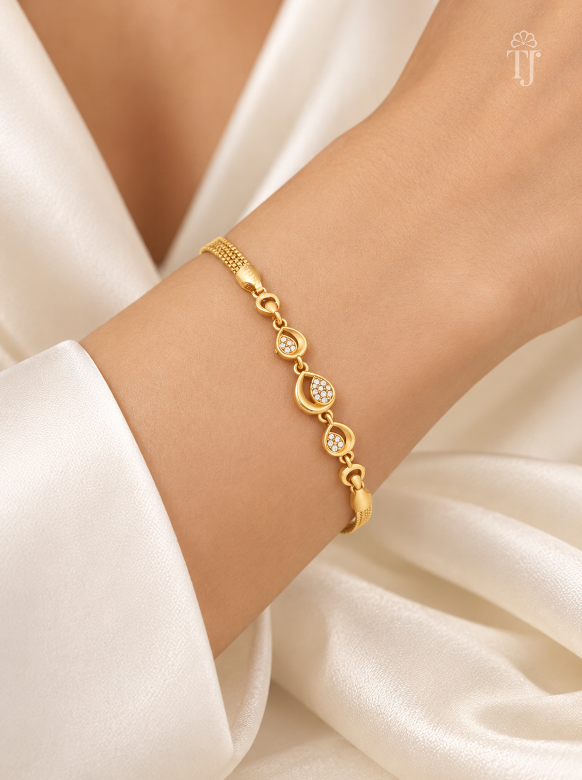 A delicate gold chain bracelet with three oval links, each set with a small clear crystal, worn on a woman's wrist. The background consists of soft, draped white satin fabric.