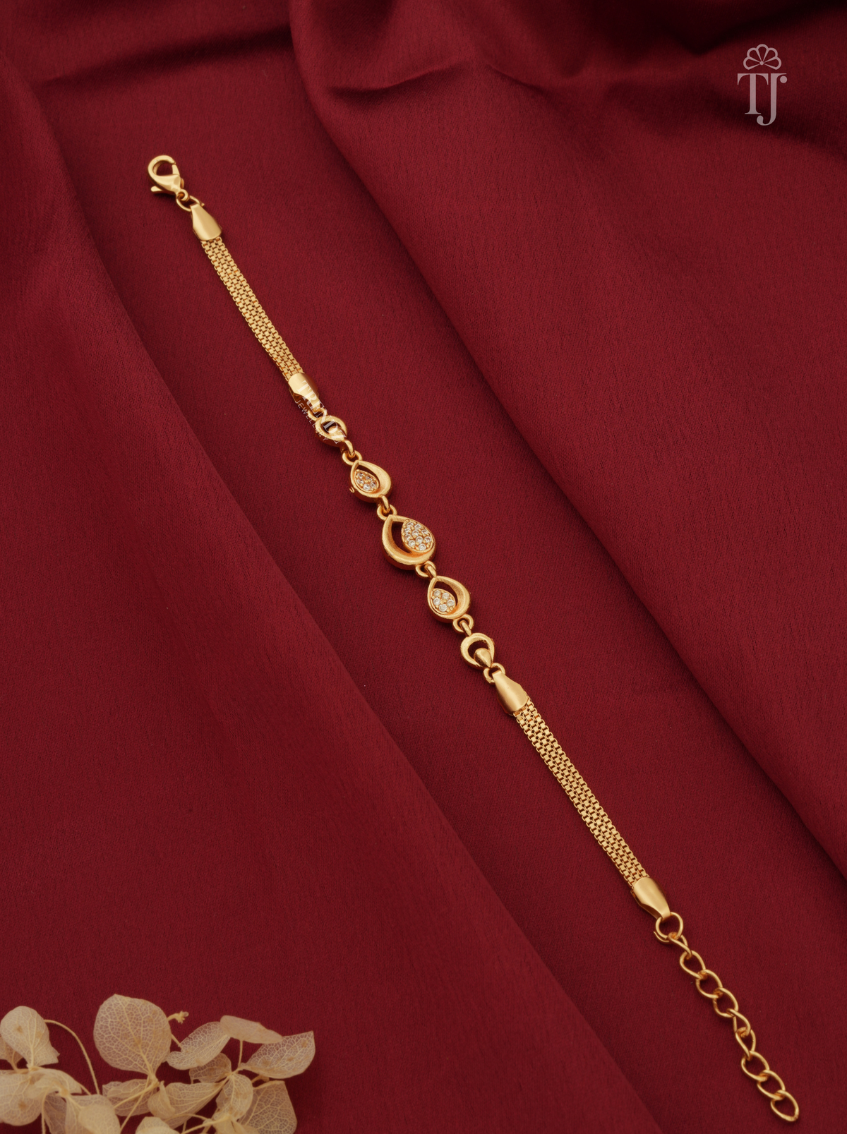 A gold-toned chain bracelet with oval and round link details, displayed diagonally on draped dark red fabric with a small dried floral accent in the corner.