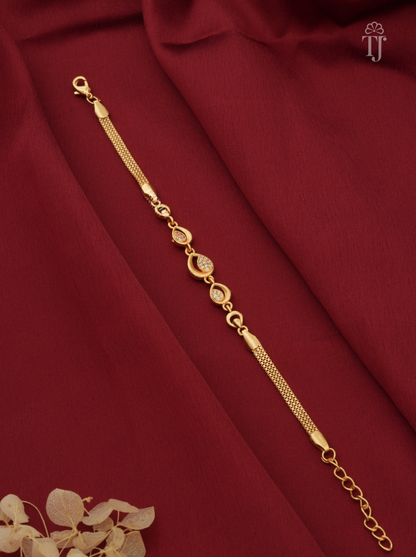A gold-toned chain bracelet with oval and round link details, displayed diagonally on draped dark red fabric with a small dried floral accent in the corner.