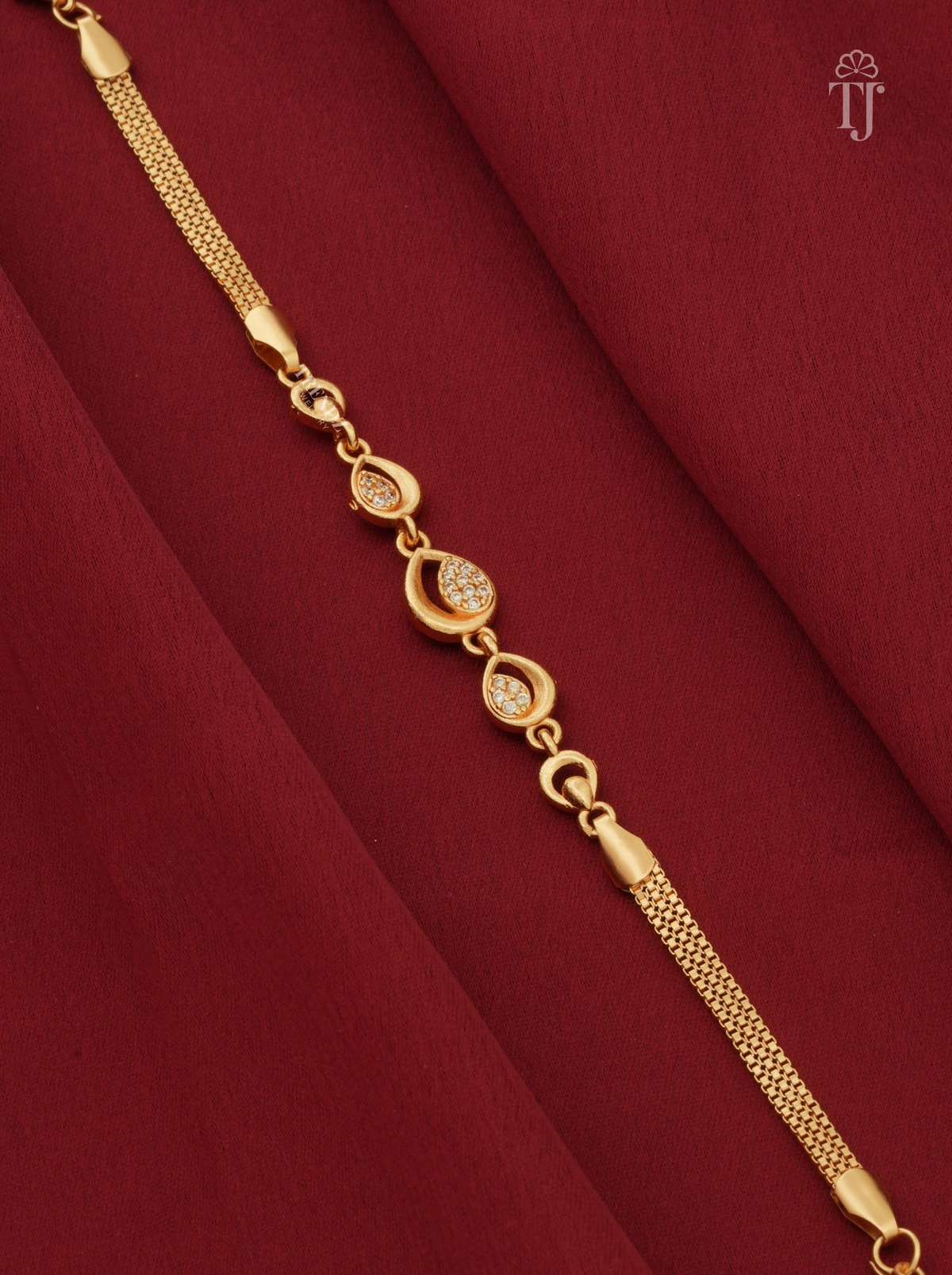 A gold chain bracelet with decorative oval links, some featuring small clear stones, displayed diagonally on a draped dark red fabric background.